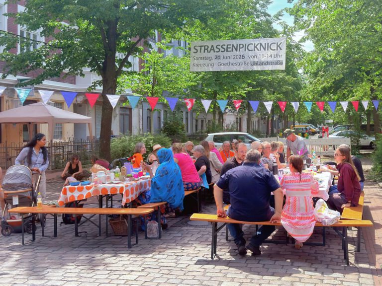 viele Menschen sitzen an Tischen auf einer Straßenkreuzung, über ihnen hängt eine bunte Wimpelkette und ein Banner auf dem 