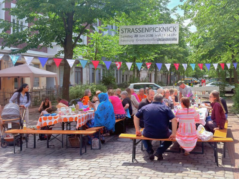 viele Menschen sitzen an Tischen auf einer Straßenkreuzung, über ihnen hängt eine bunte Wimpelkette und ein Banner auf dem 