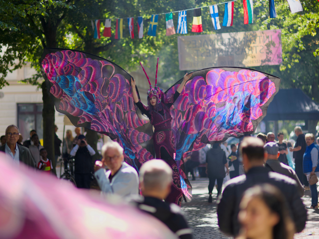 Viele Menschen auf der Goethestraße und in der mitte eine als Schmetterling verkleidete Stelzenläuferin